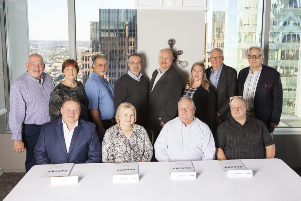 Twelve members of the Leck Waste Services management team stand in an office setting with high-rise buildings visible in the background. Among them are three women and nine men, all dressed in business attire. The group exudes professionalism and leadership, representing the diverse management team of the waste management company.
