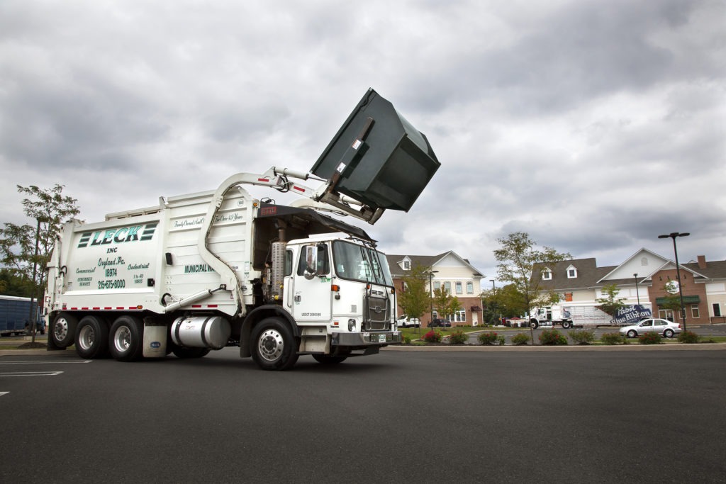 A Leck front-load truck, an Autocar model with a Heil compaction unit, is depicted in the process of emptying a dumpster into the truck's compactor. The dumpster is tilted at an angle, with its contents spilling into the opening atop the truck. The scene captures the efficient waste collection process, showcasing the specialized equipment used by Leck Waste Services for garbage disposal.